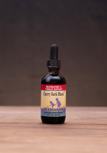 A small amber glass dropper bottle labeled Cherry Bark Blend stands centered on a wooden tabletop against a dark brown backdrop. The bottle is surrounded by rustic props, including a cork stopper, a metal tin lid, a white mortar and pestle, and a small jar, creating a styled product presentation.