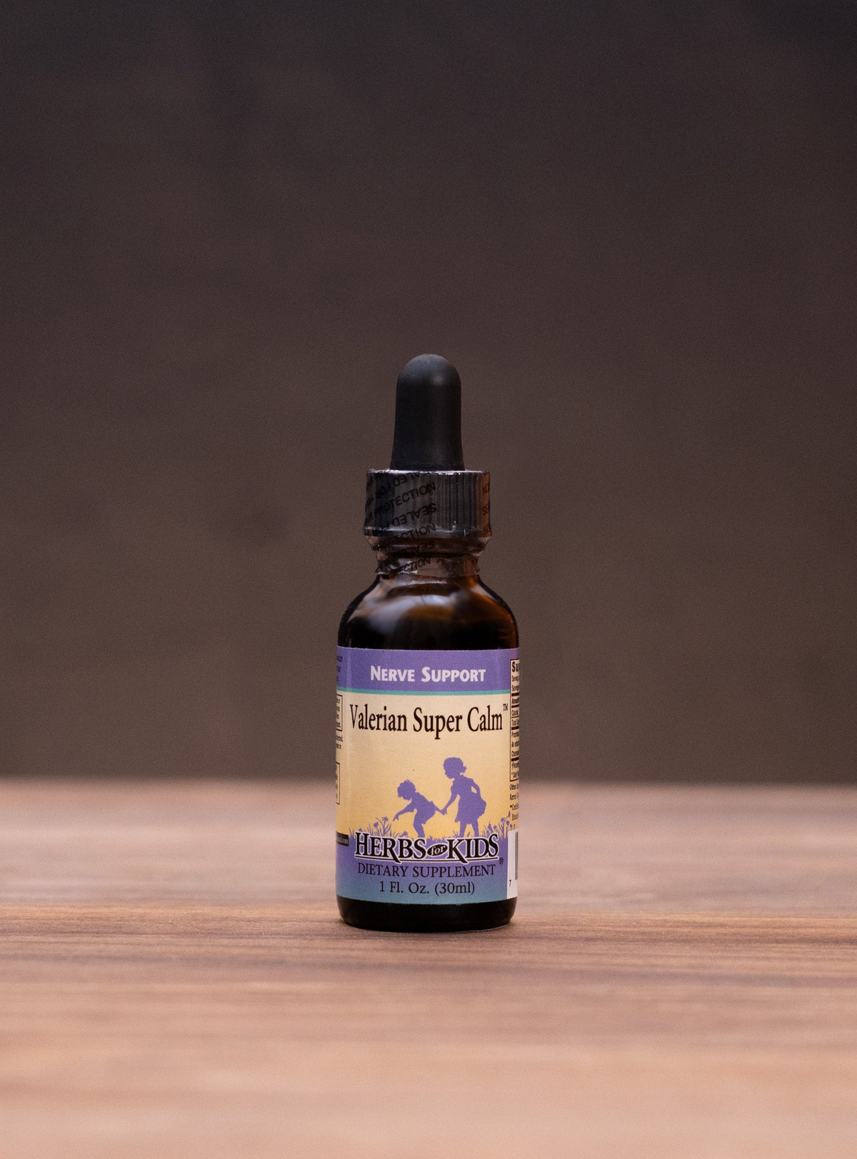 A small amber glass dropper bottle labeled Natural Super Cure stands centered on a wooden tabletop against a dark backdrop, with blurred herbal props on both sides.