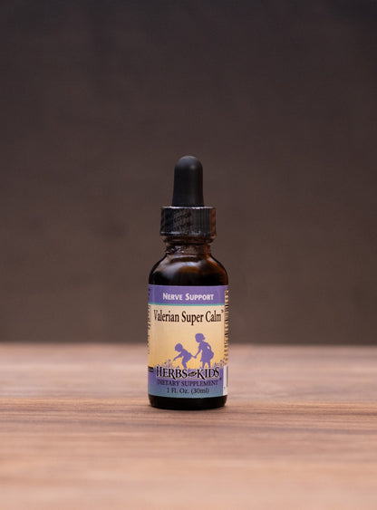 A small amber glass dropper bottle labeled Natural Super Cure stands centered on a wooden tabletop against a dark backdrop, with blurred herbal props on both sides.