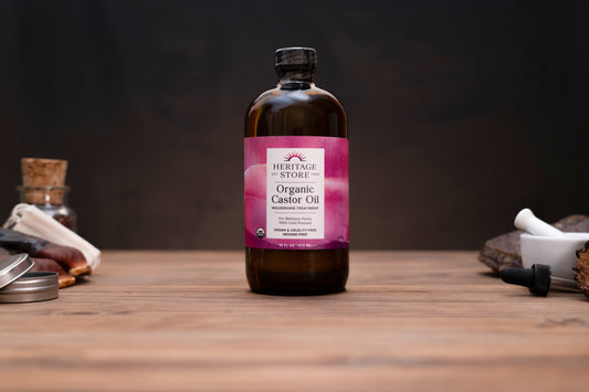 A bottle of Heritage Sturgeon Organic Castor Oil stands on a wooden tabletop against a dark background, surrounded by rustic props including a cork stopper, metal tin, white mortar and pestle, and dried herbs.