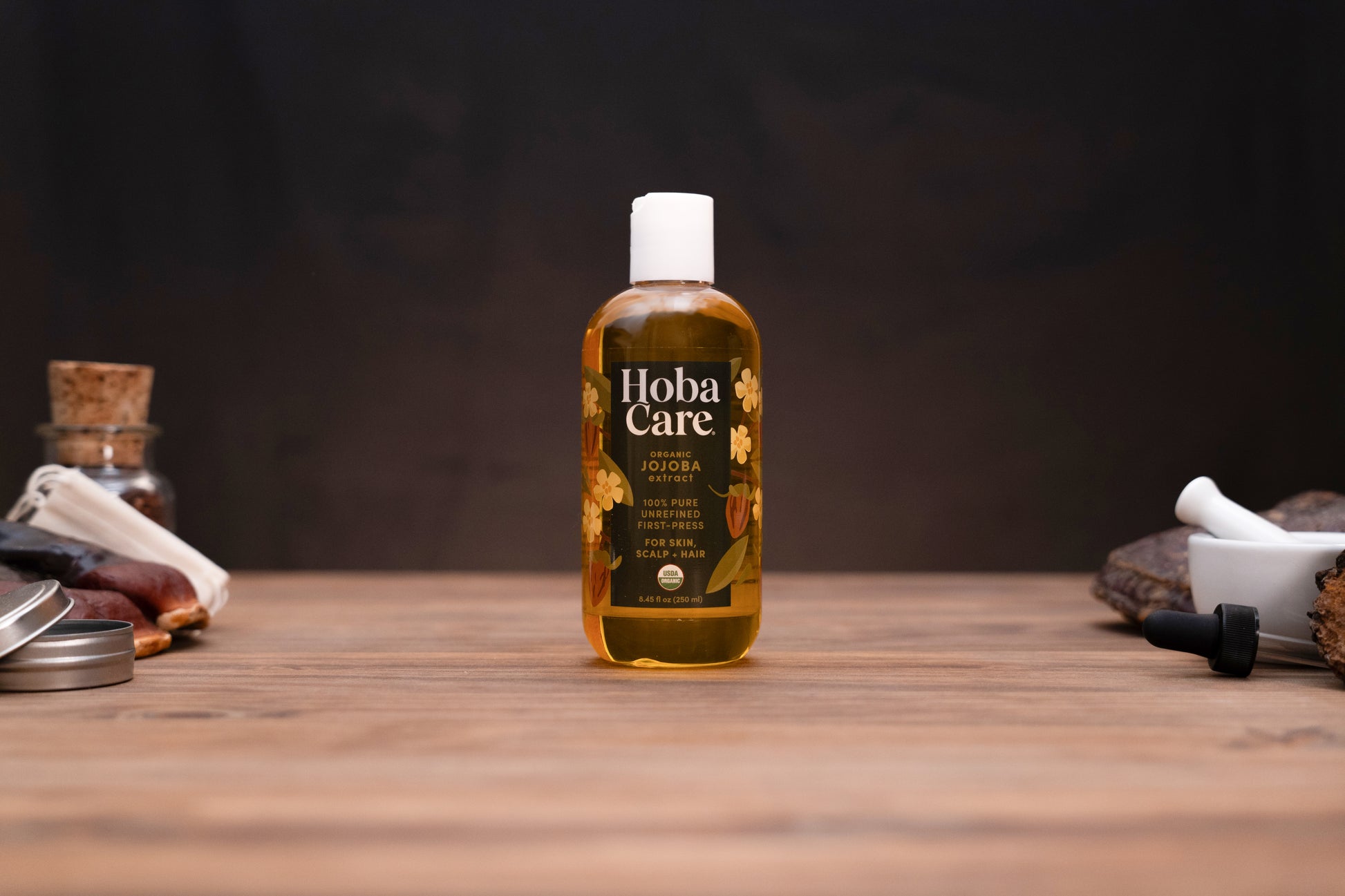 A bottle of Hoba Care Sacred Jojoba Oil stands centered on a rustic wooden table, surrounded by traditional herbal props against a dark, moody background.