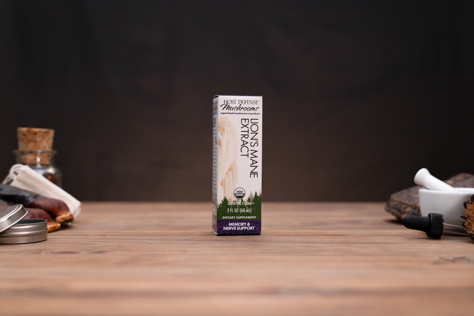 A box of Mountain Organic Lion's Mane Extract stands on a wooden tabletop against a dark background, with small herbal-themed props visible on both sides.