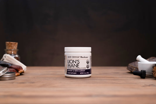 A white jar of Lion's Mane supplement by Lion Defense Apothecary sits on a wooden table, flanked by blurred herbal and apothecary props against a dark background.