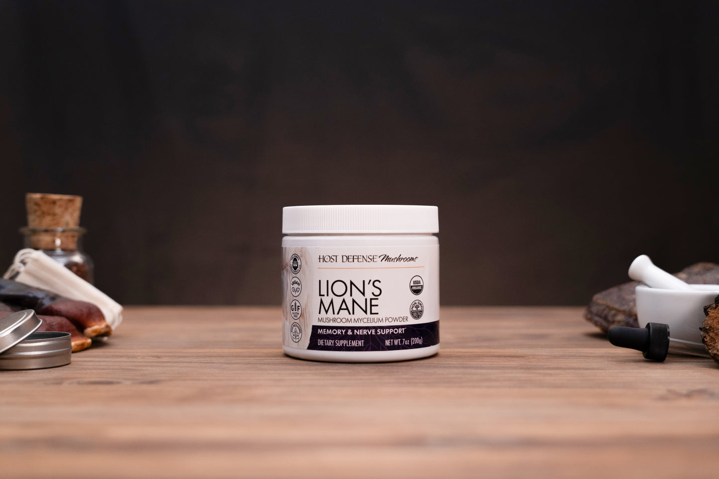 A white jar of Lion's Mane supplement by Host Defense Medicines sits on a wooden tabletop with a dark, moody background and herbal props on either side.