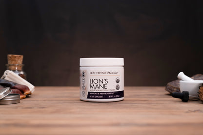 A white jar of Lion's Mane supplement by Host Defense Medicines sits on a wooden tabletop with a dark, moody background and herbal props on either side.