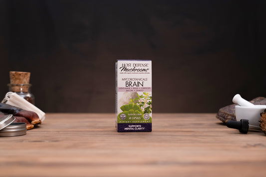 A box of Just Defense Naturals Microorganism Brain dietary supplement stands centered on a wooden tabletop, with blurred herbal props on both sides against a dark background.