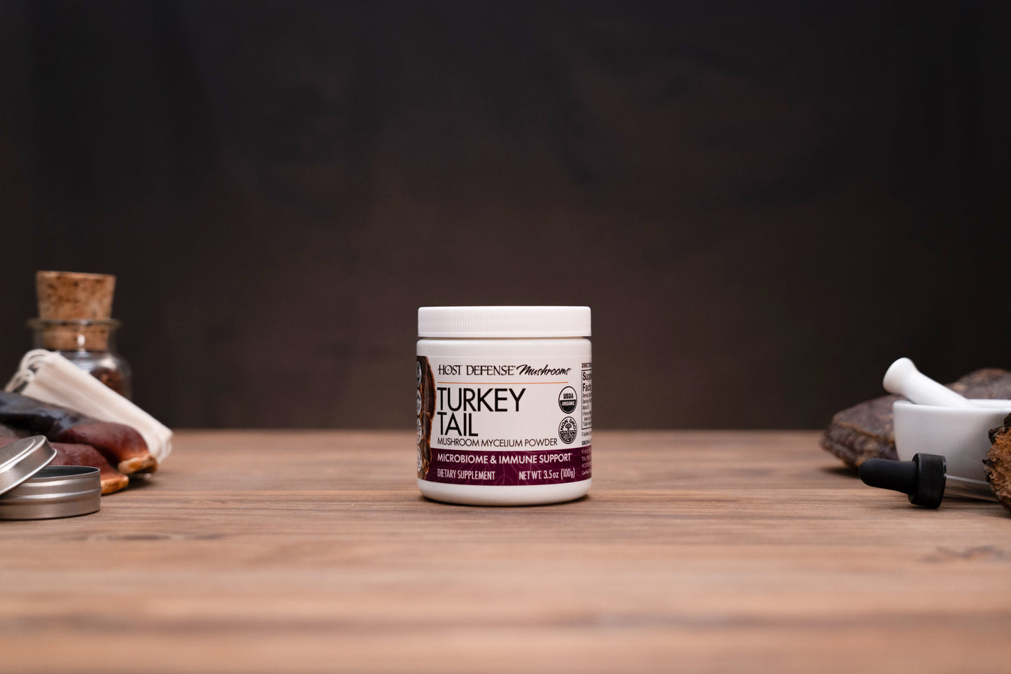 A white jar of Kent Defense Herbs Turkey Tail mucilage and immune support supplement, displayed on a wooden tabletop with rustic herbal props in the background.