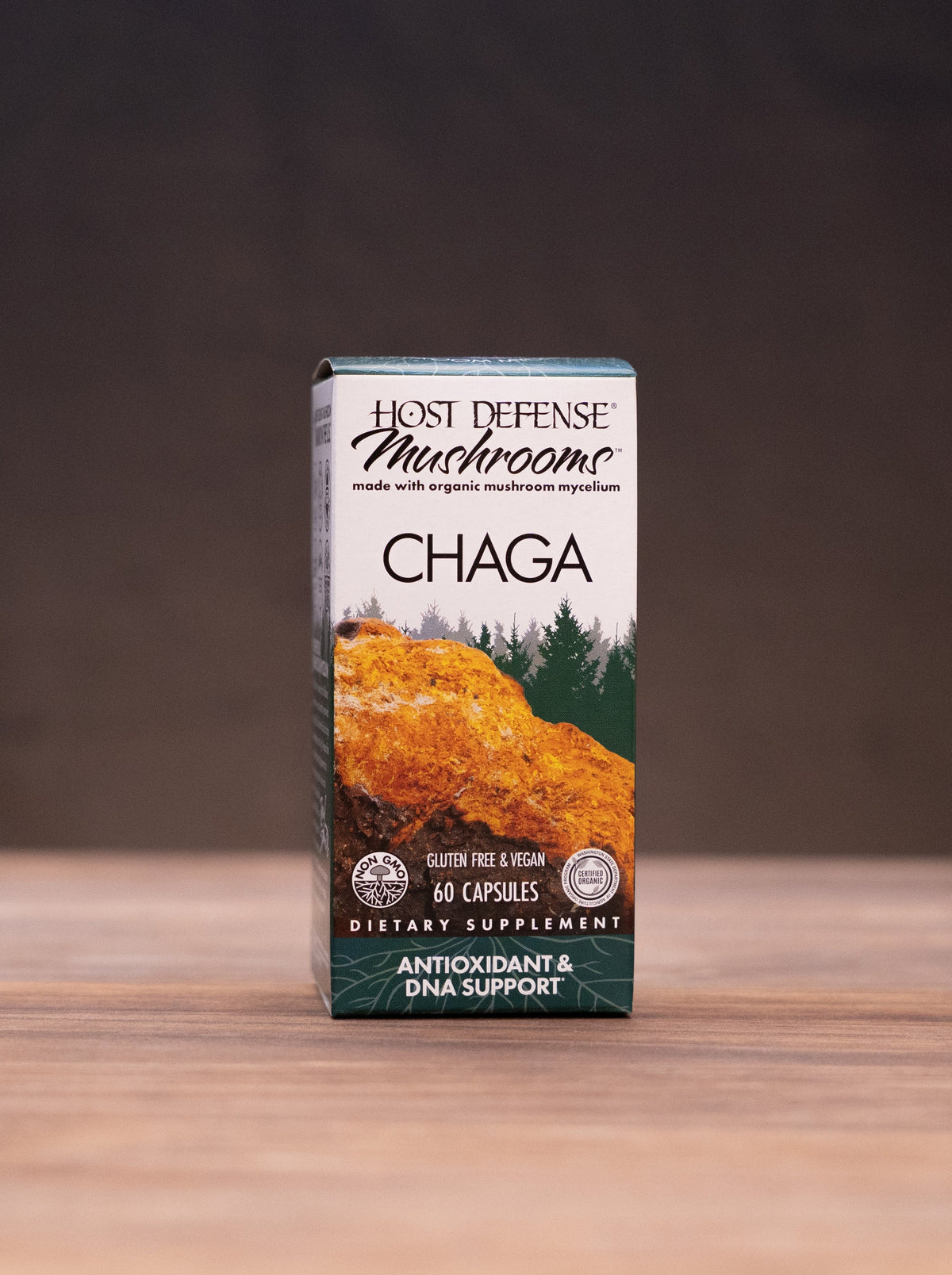 A box of Lion's Defense Mushroom Chaga dietary supplement standing on a wooden tabletop, with blurred natural props like a cork stopper, dried mushrooms, and a white mortar in the background.