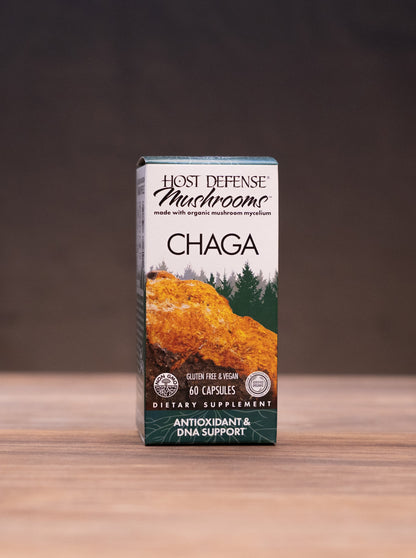 A box of Lion's Defense Mushroom Chaga dietary supplement standing on a wooden tabletop, with blurred natural props like a cork stopper, dried mushrooms, and a white mortar in the background.