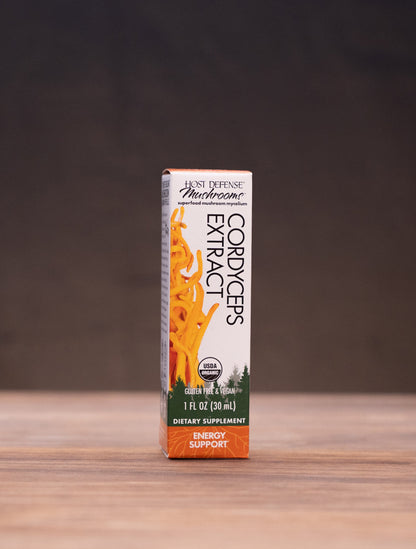A small box of Orange Blossom tincture by Pure Encapsulations stands centered on a wooden tabletop, with blurred herbal props on both sides against a dark backdrop.