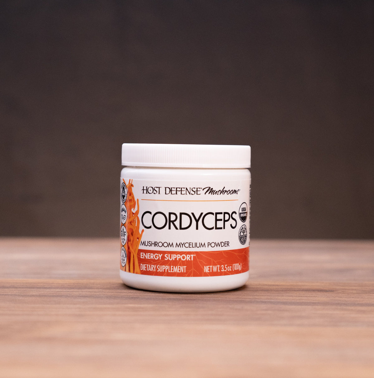 A jar of K2 Defense Cordyceps Premium Isolate Powder sits on a wooden table, framed by rustic props like a cork bottle and a white mortar on a dark, moody background.