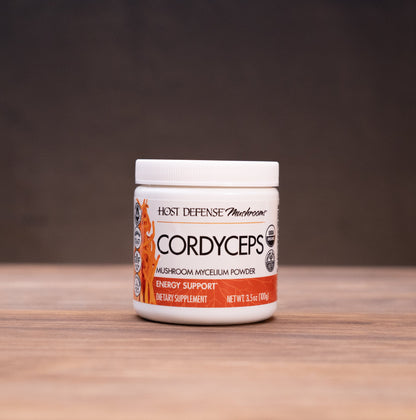 A jar of K2 Defense Cordyceps Premium Isolate Powder sits on a wooden table, framed by rustic props like a cork bottle and a white mortar on a dark, moody background.