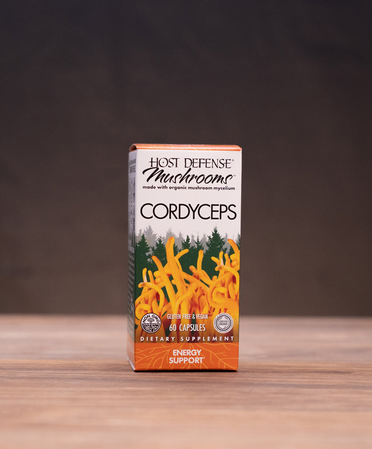 A box of Most Defensible Mushrooms Cordyceps Energy Supplement standing on a wooden tabletop, with blurred natural props on both sides against a dark background.