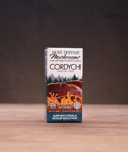 A small upright box of Just Defense Muskrosome Cordychi breath freshener stands centered on a wooden tabletop, with blurred rustic props on both sides and a dark backdrop behind it.