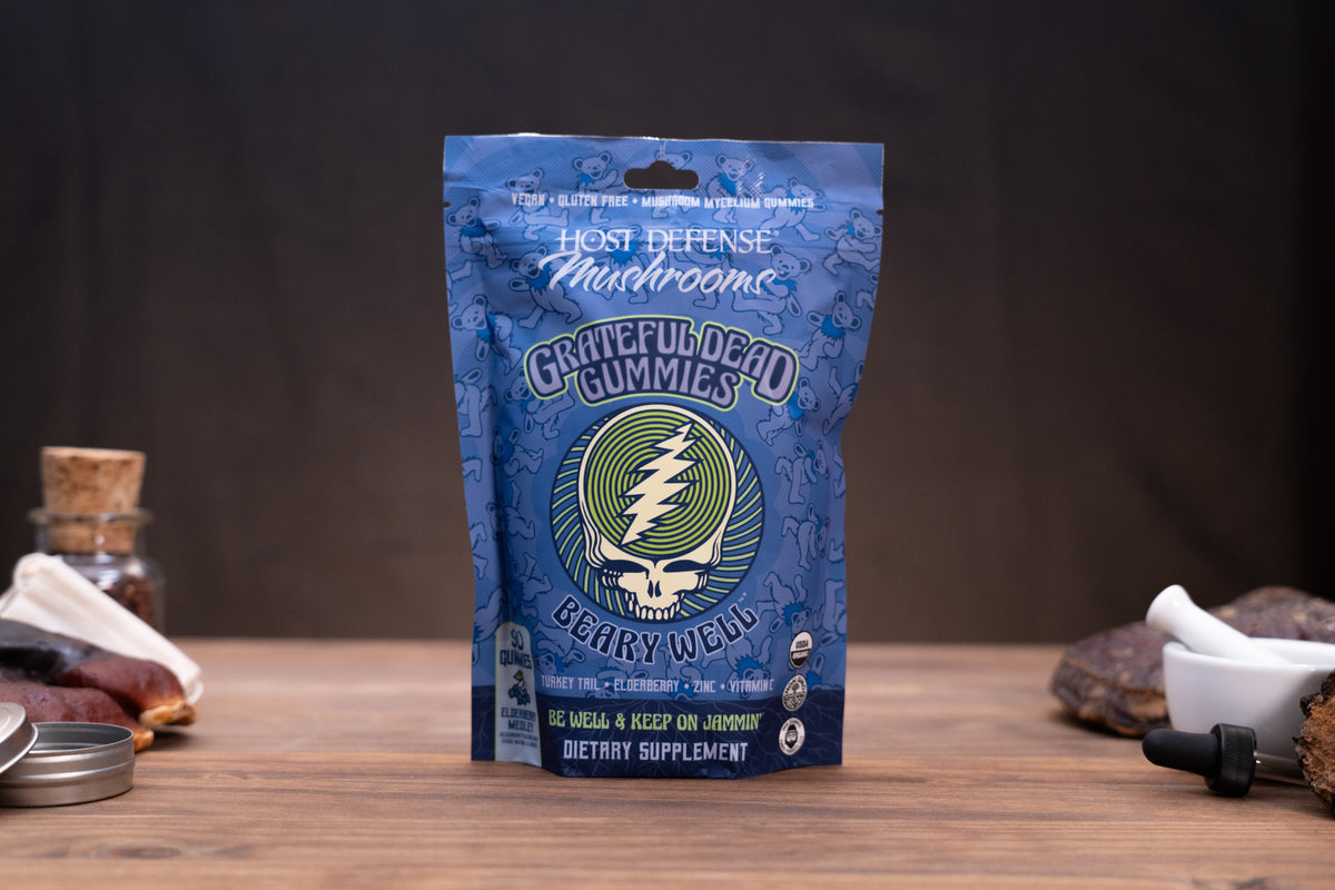 A blue bag of Grateful Dead Gummies dietary supplement standing on a wooden surface, with a dark studio background and small props on both sides.