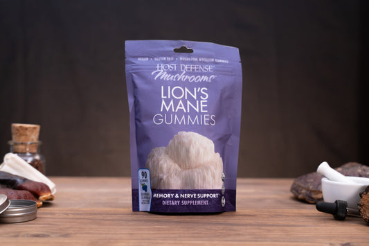 A purple stand-up pouch of Host Defense Mushrooms Lion's Mane Gummies stands on a wooden surface against a dark background, with small herbal and kitchen props blurred around it.
