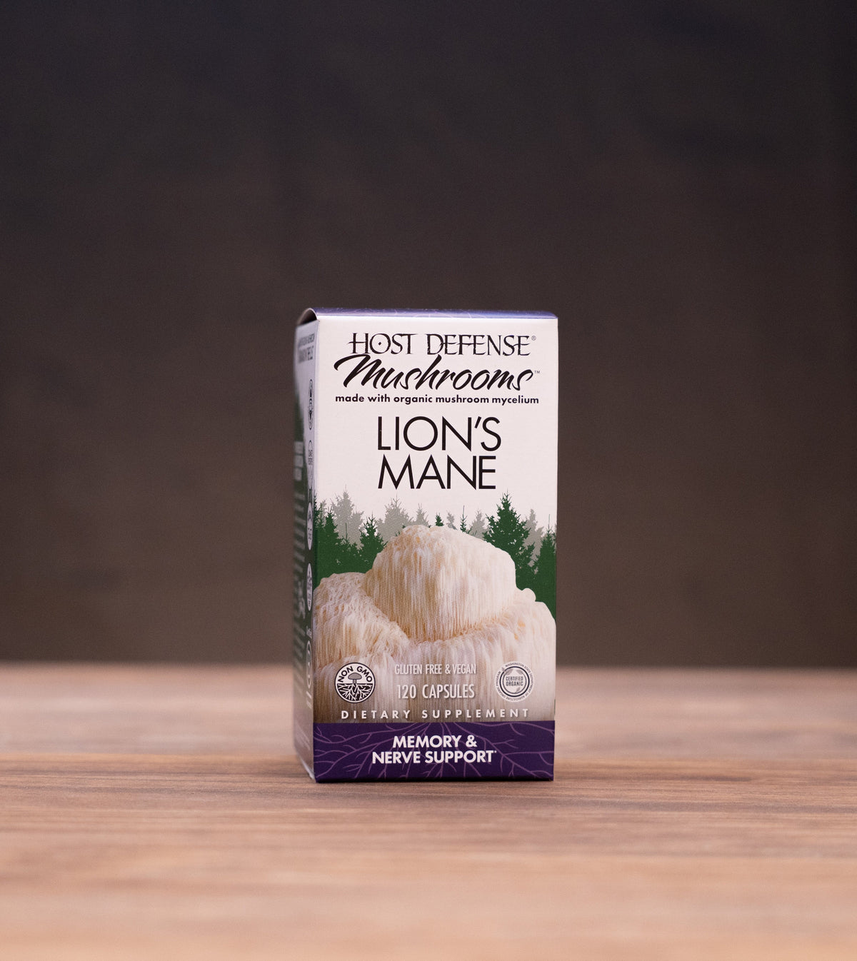 A box of Host Defense Mushroom Lion's Mane supplement stands centered on a wooden tabletop, with rustic props softly blurred in the background and foreground.