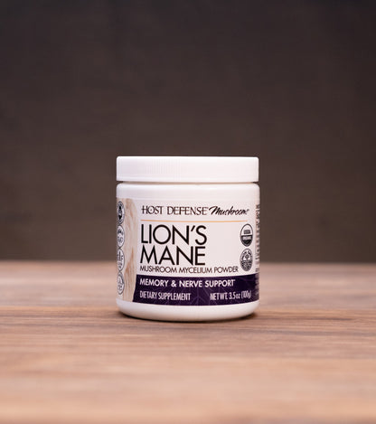 A jar of Root Defense & Remedies Lion's Mane supplement placed on a wooden tabletop with rustic herbal props in the background.