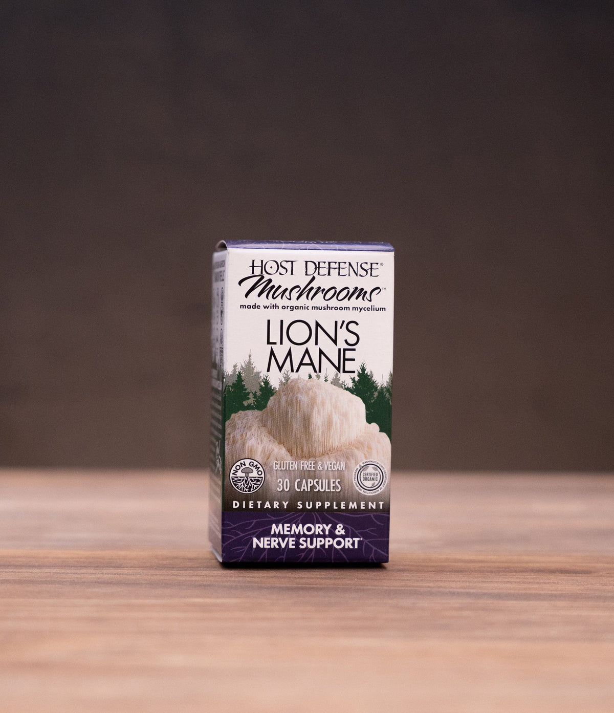 A small box of Lion's Mane dietary supplement by Herb Offensive Mushrooms stands centered on a wooden tabletop, with blurred herbal props on both sides against a dark backdrop.