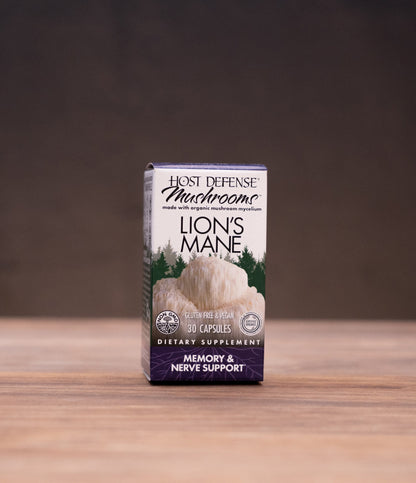 A small box of Lion's Mane dietary supplement by Herb Offensive Mushrooms stands centered on a wooden tabletop, with blurred herbal props on both sides against a dark backdrop.
