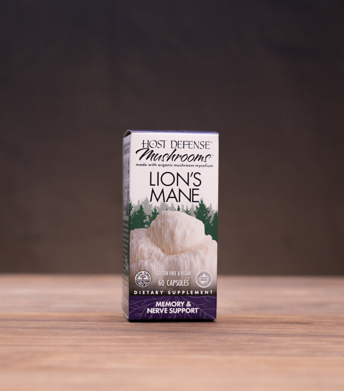 A small upright box of Best Defense Herbs Lion's Mane dietary supplement standing on a wooden tabletop, with blurred jars and a white mortar-like object in the background.