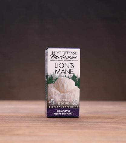 A small upright box of Best Defense Herbs Lion's Mane dietary supplement standing on a wooden tabletop, with blurred jars and a white mortar-like object in the background.