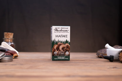 A box of Host Defense Mushroom Co. Maitake dietary supplement capsules stands centered on a wooden tabletop, with blurred jars, stones, and a white mortar in the background.