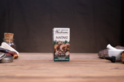 A box of Maitake mushroom powder by First Defense Mushrooms stands centered on a wooden tabletop, with blurred rustic props on both sides against a dark backdrop.