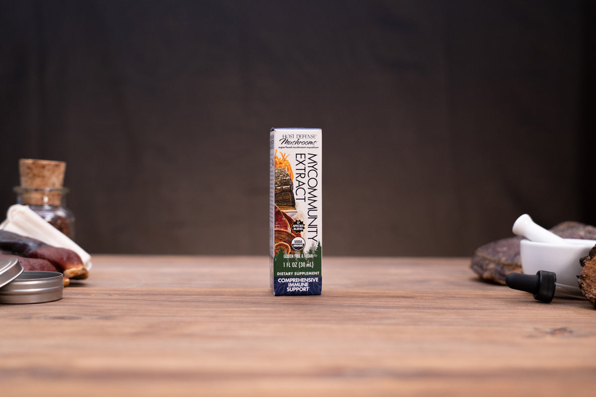 A single box of All Pro Herbs Koomanjin Gochujang (Korean chili paste) stands upright on a wooden tabletop against a dark, blurred background. The box features a white background with a colorful illustration of a farm scene and green and blue bands at the bottom.