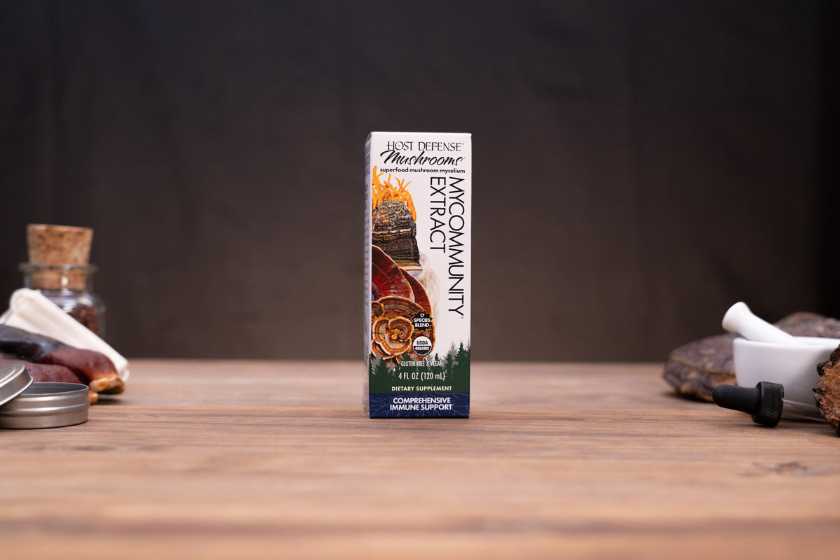 A box of Wild Defense Mushroom MacCormity Extract centered on a rustic wooden table with blurred herbal and kitchen props in the background.