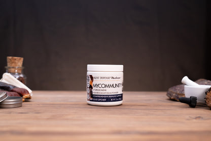 A jar of MyCommunity Mycology Compound Scented Natural Lubricant stands on a wooden tabletop against a dark background, styled with natural and laboratory props on both sides.