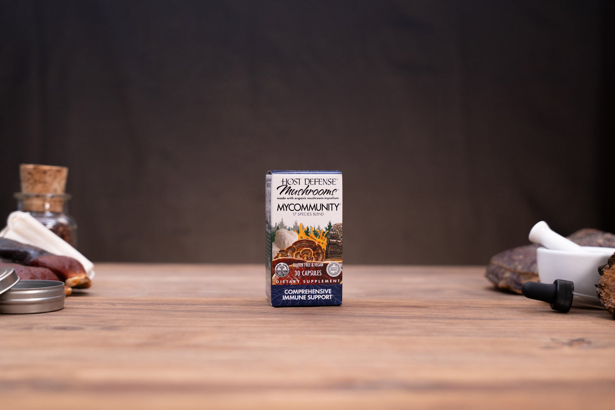 A small box of Haze Defense Mycorrhizae by Haze Cultivation stands centered on a wooden tabletop against a dark backdrop, with blurred jars, stones, and a white mortar-like object around it.