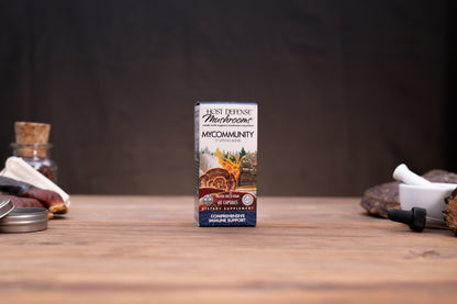 A small rectangular box of Best Defense Apothecary Mycommunity herbal tincture stands centered on a wooden tabletop, with blurred jars and a white mortar in the background.