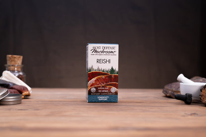 A box of Host Defense Mushroom Co Reishi powder stands centered on a wooden tabletop against a dark background, with small rustic props visible at the left and right edges.