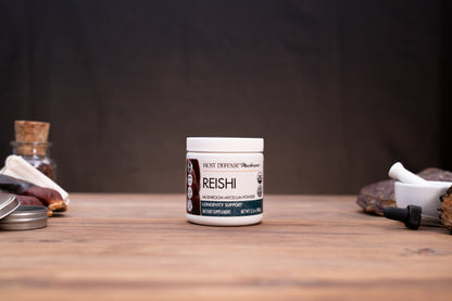 A white jar of REISHI Root Defense Hair Growth Support centered on a wooden tabletop, with blurred natural props on both sides against a dark backdrop.