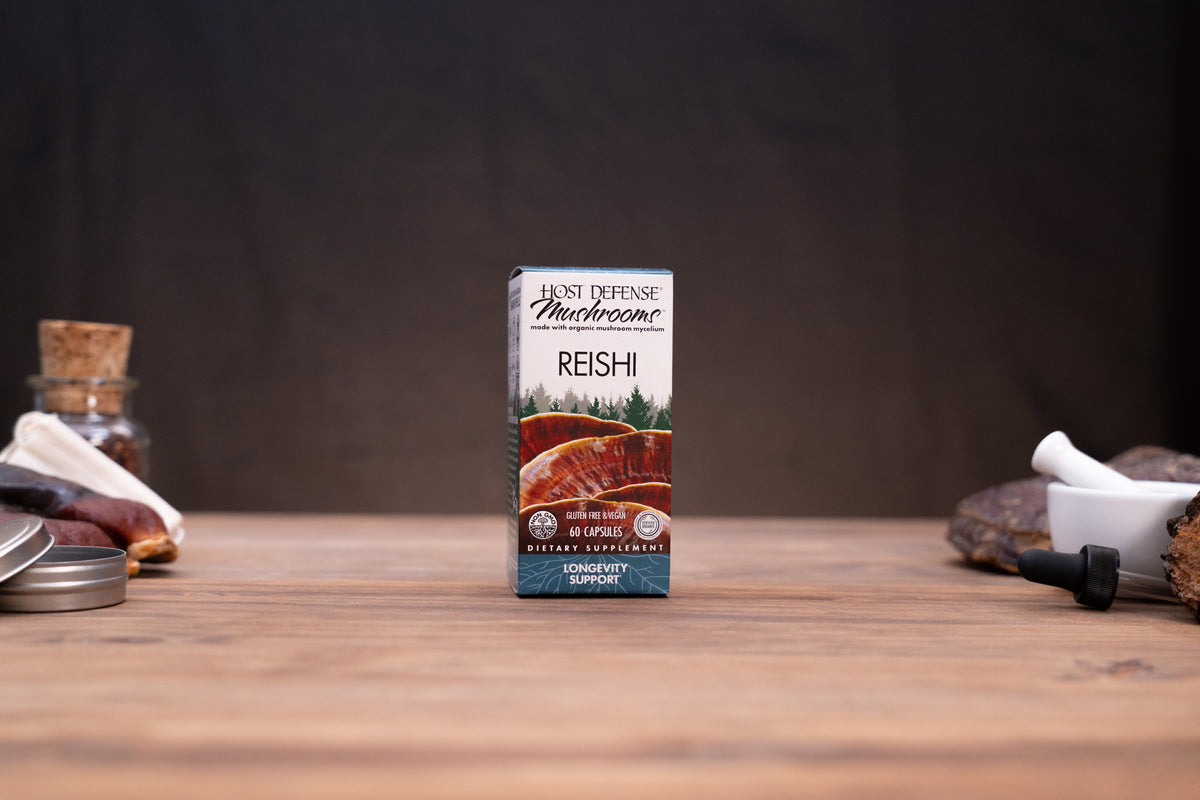 A box of Host Defense Microbes Reishi Conevity Support capsules stands centered on a wooden tabletop, with blurred rustic props on both sides against a dark backdrop.
