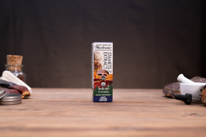 A box of Pine Ridge Apothecary Energy Extrait coffee concentrate stands centered on a wooden tabletop, with blurred rustic props on both sides against a dark background.