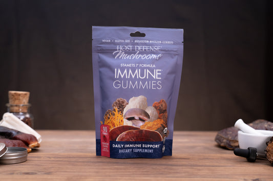 A stand-up pouch of Host Defense Mushrooms Immune Gummies displayed on a wooden surface with small kitchen-style props around it against a dark background.
