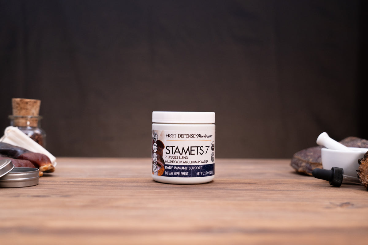 A jar of Hot Defense Medicinals Stamets 7 protein powder sits on a wooden table, surrounded by blurred ingredients and kitchen props in the background.