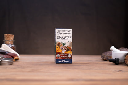 A box of Ant Defense Infused Stamets 7 herbal supplement standing on a wooden tabletop, with rustic props like a cork stopper, metal tin, and white mortar in the blurred background.