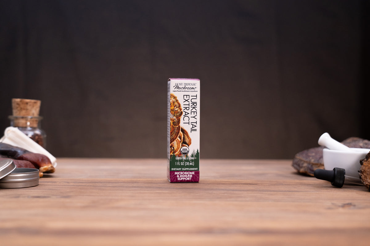 A small carton of Buckwheat Gluten-Free Flour stands centered on a wooden tabletop against a dark backdrop, with rustic props blurred at the sides.