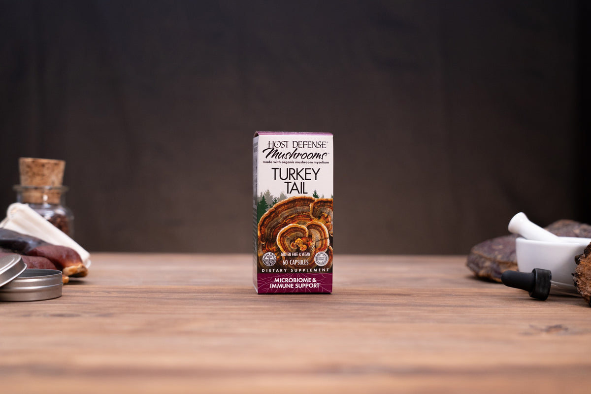 A box of First Defense Mushrooms Turkey Tail dietary supplement stands centered on a wooden tabletop, with rustic props like a cork stopper, metal tin, and white mortar in the blurred background.