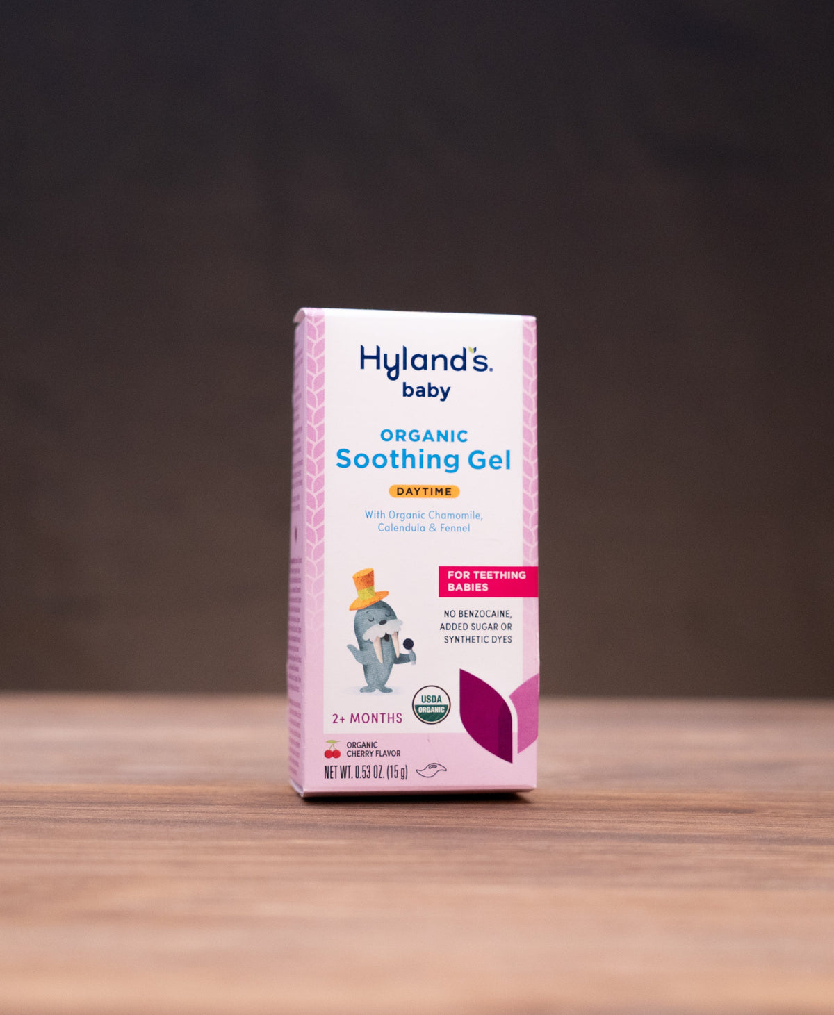 A box of Hyland's Baby Organic Soothing Gel stands centered on a wooden tabletop against a dark background, with blurred natural props on both sides.