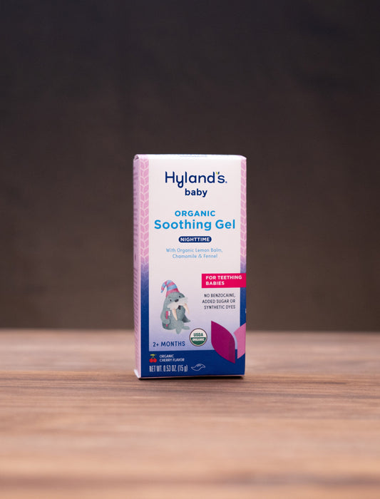 A box of Hyland's Baby Organic Soothing Gel stands centered on a wooden tabletop against a dark backdrop, with small rustic props visible on both sides.