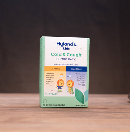 A box of Hyland's Kids Cold & Cough Combo Pack stands on a wooden tabletop against a dark background, with small jars and a mortar-and-pestle props placed on both sides.