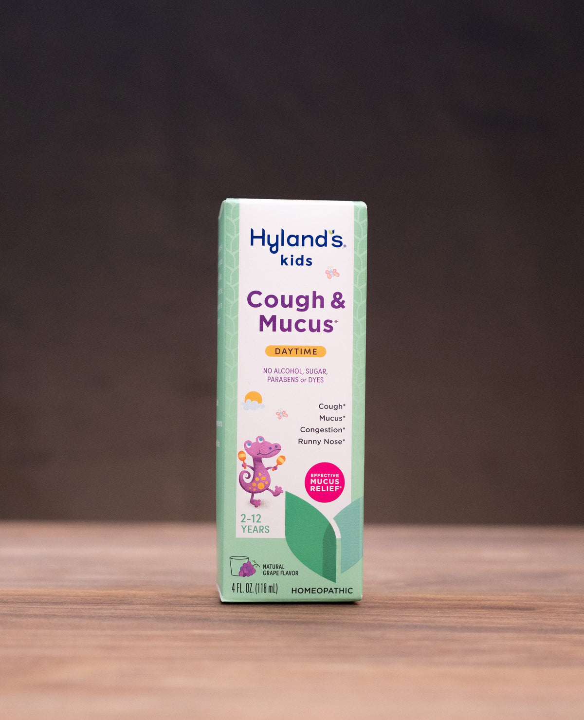 A box of Hyland's Kids Cough & Mucus syrup stands centered on a wooden tabletop against a dark backdrop, with blurred natural props on both sides.