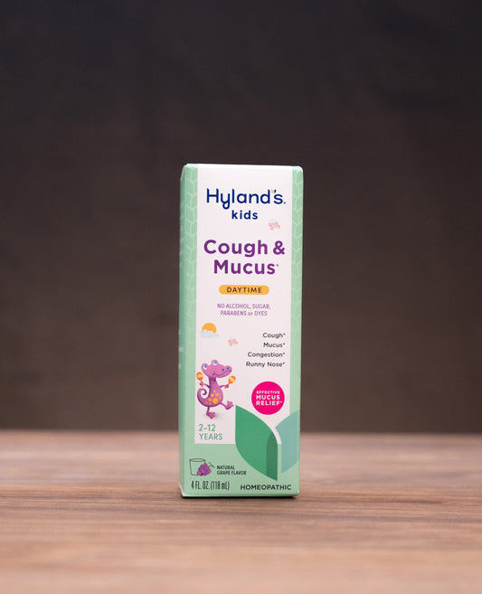 A box of Hyland's Kids Cough & Mucus syrup stands centered on a wooden tabletop against a dark backdrop, with blurred natural props on both sides.