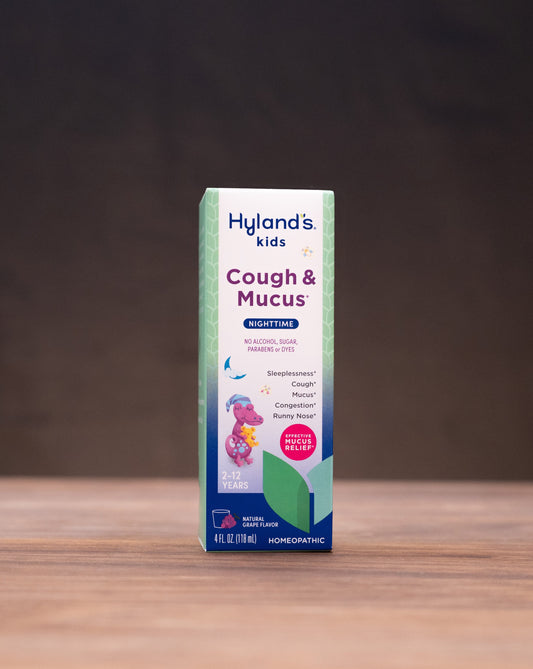 A box of Hyland's Kids Cough & Mucus syrup stands centered on a wooden tabletop against a dark brown background, with small blurred props on both sides.