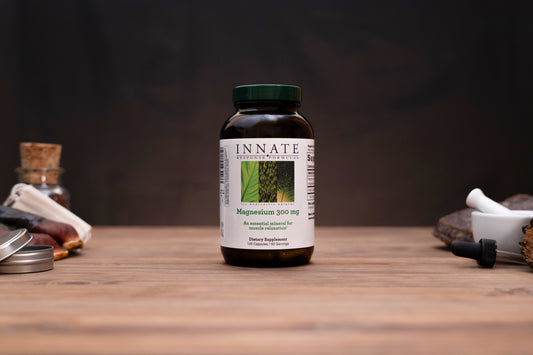 A bottle of Innate Botanicals Magnesium 310 mg dietary supplement, set on a wooden table with blurred herbal props in the background.