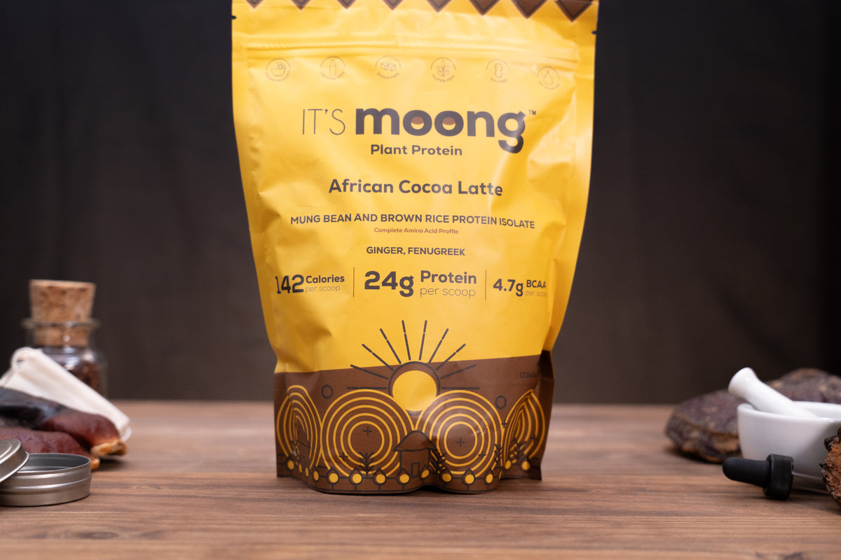 A yellow stand-up pouch of It's Moong Plant Protein African Cocoa Latte stands on a wooden surface against a dark background, with small ingredients and tools placed around it.